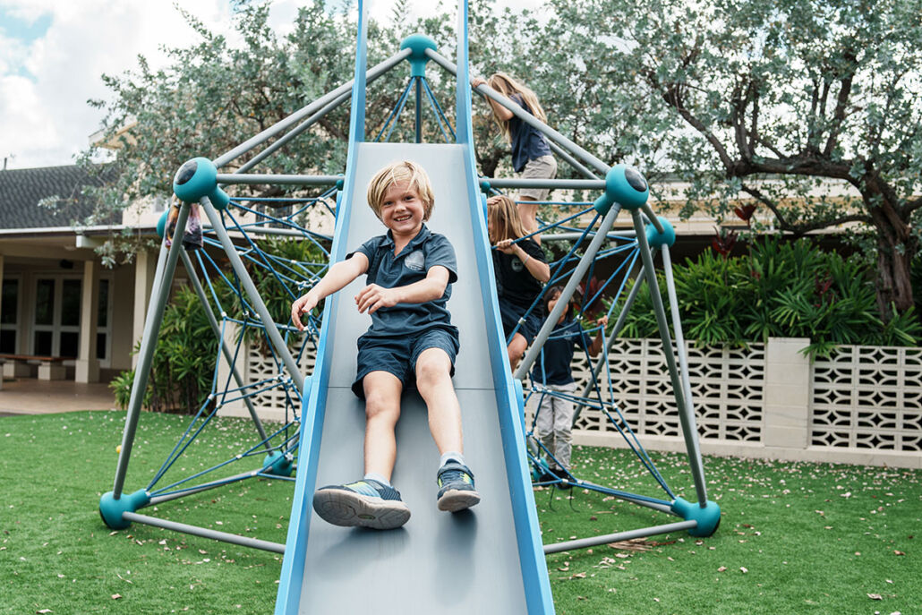Smiling child sliding down playground structure onto clean artificial turf maintained with TurFresh for safe school play areas.