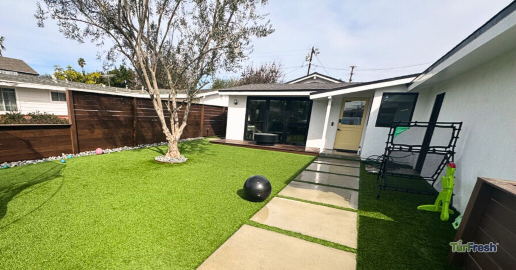 Clean backyard artificial turf lawn surrounding a tree and patio area, professionally maintained by TurFresh for a lush, green appearance.
