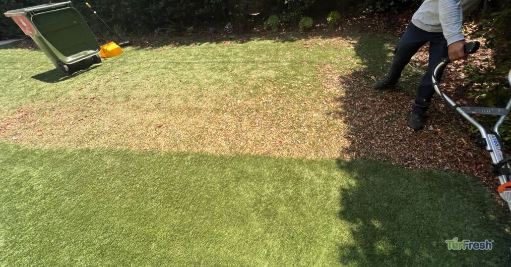 Technician using a power broom to clean debris from worn artificial turf lawn, serviced by TurFresh.