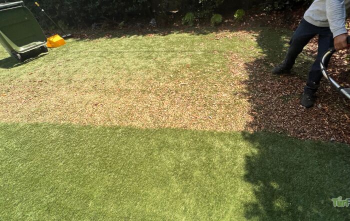 Technician using a power broom to clean debris from worn artificial turf lawn, serviced by TurFresh.